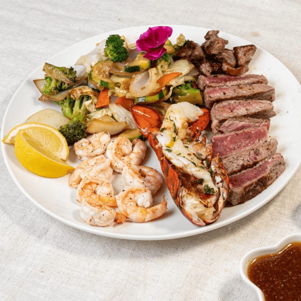 A surf and turf platter with steak, lobster, shrimp, and mixed vegetables, garnished with a lemon wedge.