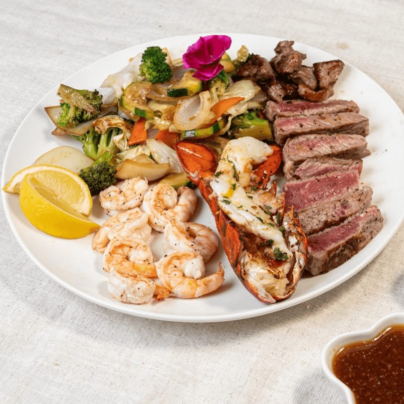 A surf and turf platter with steak, lobster, shrimp, and mixed vegetables, garnished with a lemon wedge.