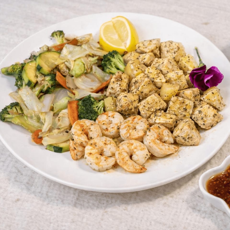 A white plate with grilled shrimp, chicken, and mixed vegetables, garnished with a lemon wedge and purple flower.